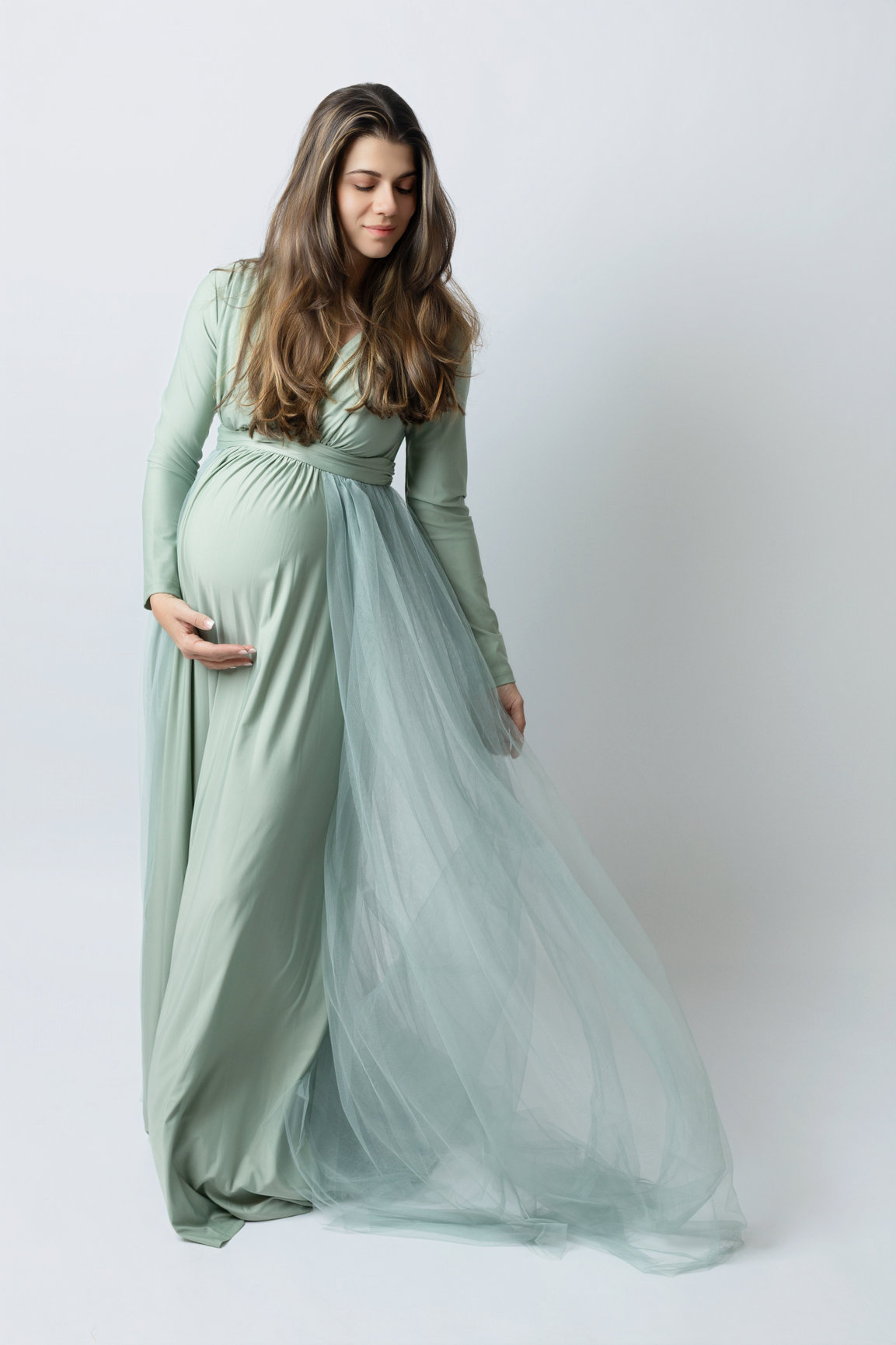 Expectant mother in a flowing sage maternity gown against a light studio background.