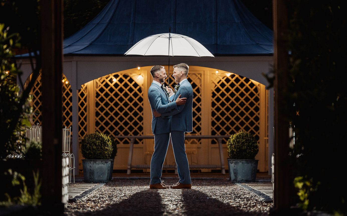 Two men in blue suits embrace under a white umbrella at a warmly lit outdoor wedding venue.