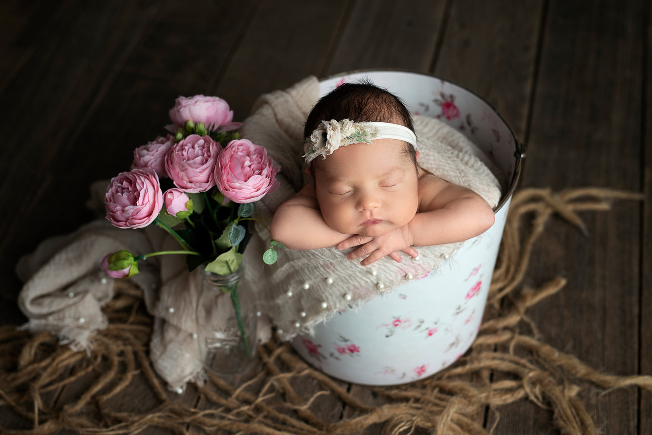In Rolling Meadows, Illinois, a sleeping baby rests in a floral bucket, wearing a white headband with a small flower. The bucket is draped with a light cloth and surrounded by pink peonies in a glass vase on the wooden floor.