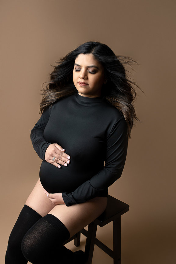 In a serene maternity photoshoot, a pregnant woman wearing a long-sleeve black outfit sits on a stool against a beige background. Her hands rest gently on her belly, eyes closed, with her hair flowing back as if caught by the wind.