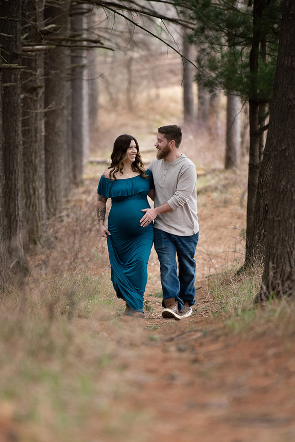 A pregnant woman in a blue dress walks hand in hand with a man through a forest, one of the best places to take photos near me. They are smiling at each other, surrounded by tall trees and stunning natural scenery.
