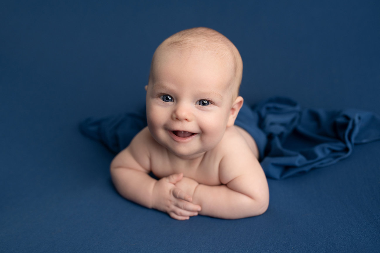 A smiling baby lies on their stomach on a soft, dark blue surface, partially covered by matching fabric. The joyful infant gazes directly at the camera, capturing the essence of newborn photography near me.