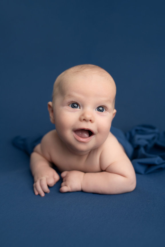 A smiling baby lies on its stomach against a dark blue background, embodying the joy of maternity photography. Wrapped in a blue cloth around its lower body, the baby appears joyful with wide eyes and an open mouth.