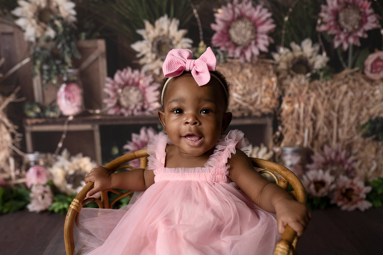 A smiling baby wearing a pink dress and headband sits on a wooden chair, captured in a charming studio photography session. The background features large pink and white flowers, hay, and wooden crates, creating a rustic, floral setting.
