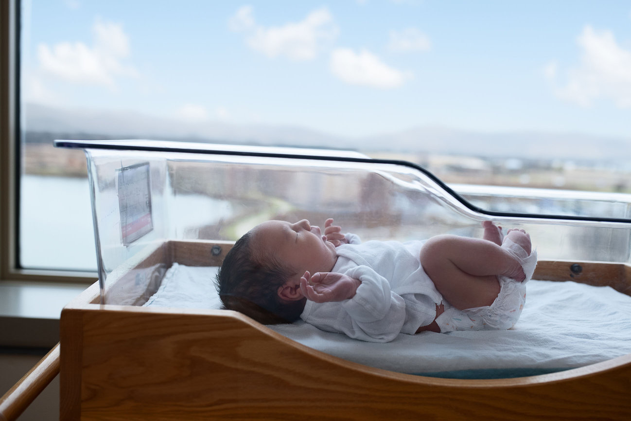 A newborn baby peacefully sleeping in a hospital bassinet at Advocate Good Shepherd Hospital in Barrington. The bassinet, crafted from clear plastic with a wooden frame, sits by a large window showcasing a scenic view.