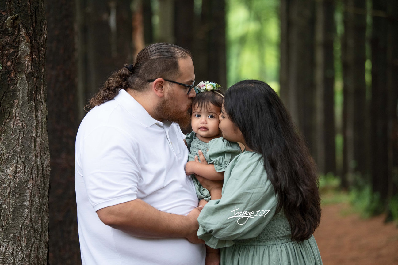 a couple lovingly kisses their baby on the cheeks, capturing a perfect family photo. The man wears a white shirt, while the green outfits that blend harmoniously with nature. The babys flower headband against the backdrop of tall trees.
