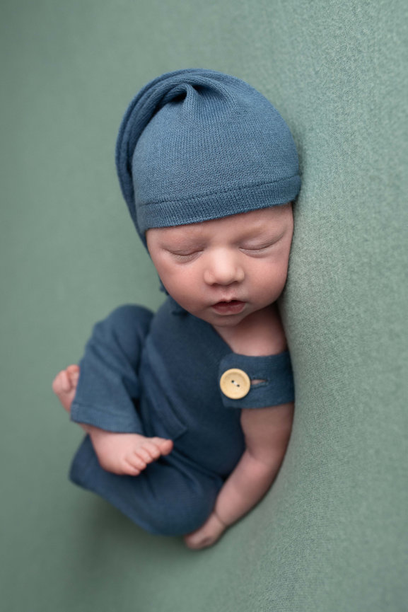 A sleeping bebé dressed in a blue outfit and matching hat, nestled on a soft green surface. The baby has a peaceful expression and is gently curled, with a wooden button on the outfit near the shoulder.