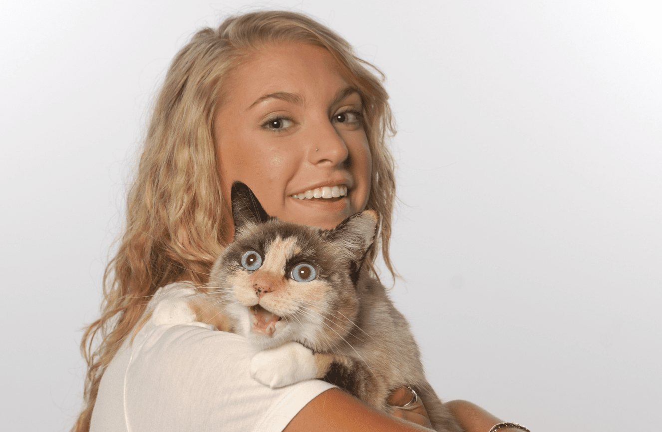 A high school blonde senior girl with her calico cat over her shoulder meowing for his life in the Kliks Photography studio.
