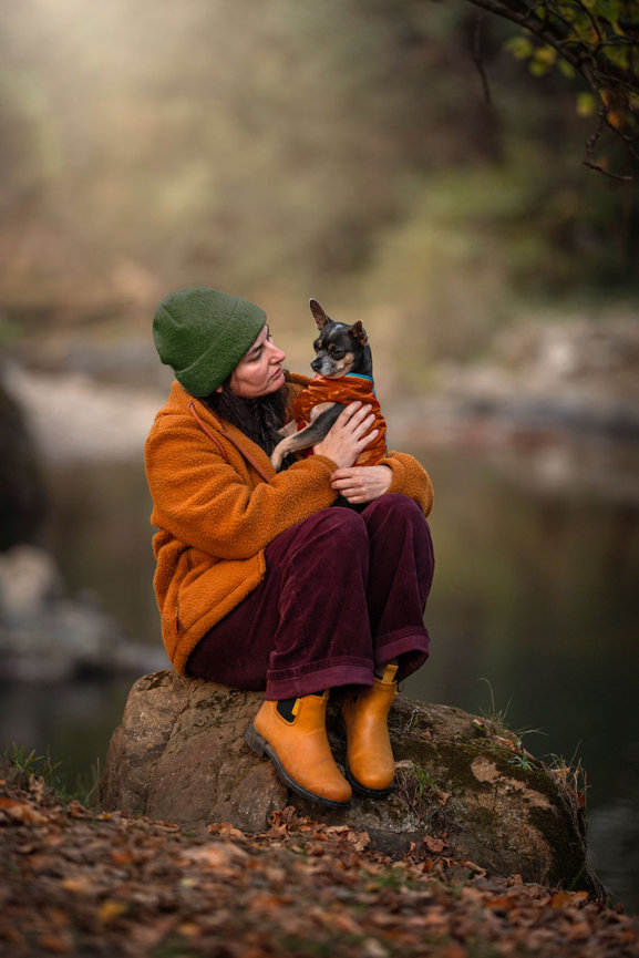 A person in a green beanie and orange jacket sitting on a rock beside a river, holding a small chihuahua wearing an orange jumper
