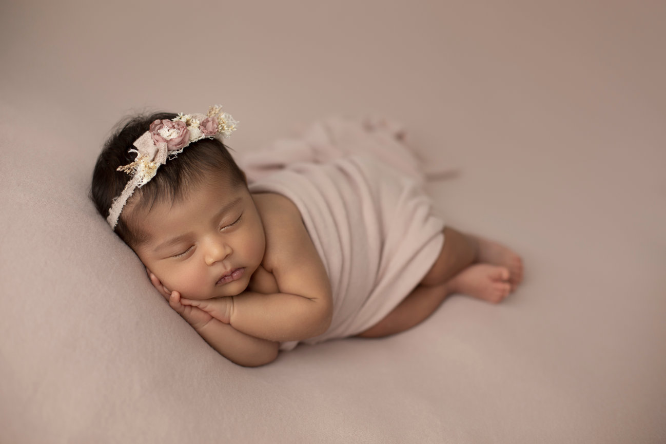 Dark skinned baby girl on mauve backdrop with matching wrap and headband, posed on her side with hands sweetly tucked under her cheek.