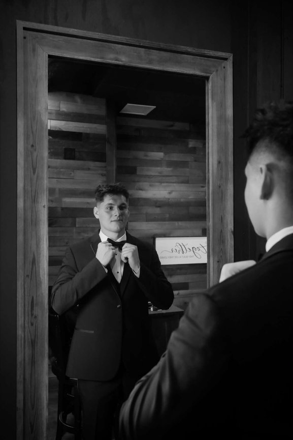 Groom adjusting his bow tie while looking in a mirror.