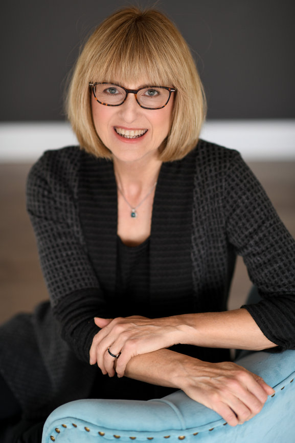 A smiling woman with blonde hair and glasses seated on a blue chair against a dark background. Professional Headshots in Columbus Ohio