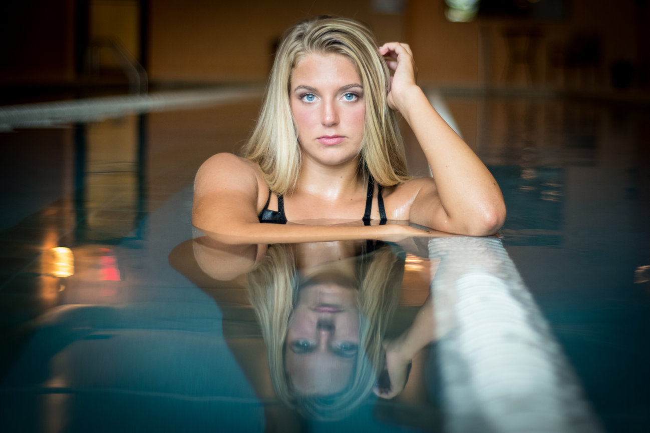 A young woman with blonde hair poses thoughtfully in a swimming pool, her reflection visible on the water's surface.