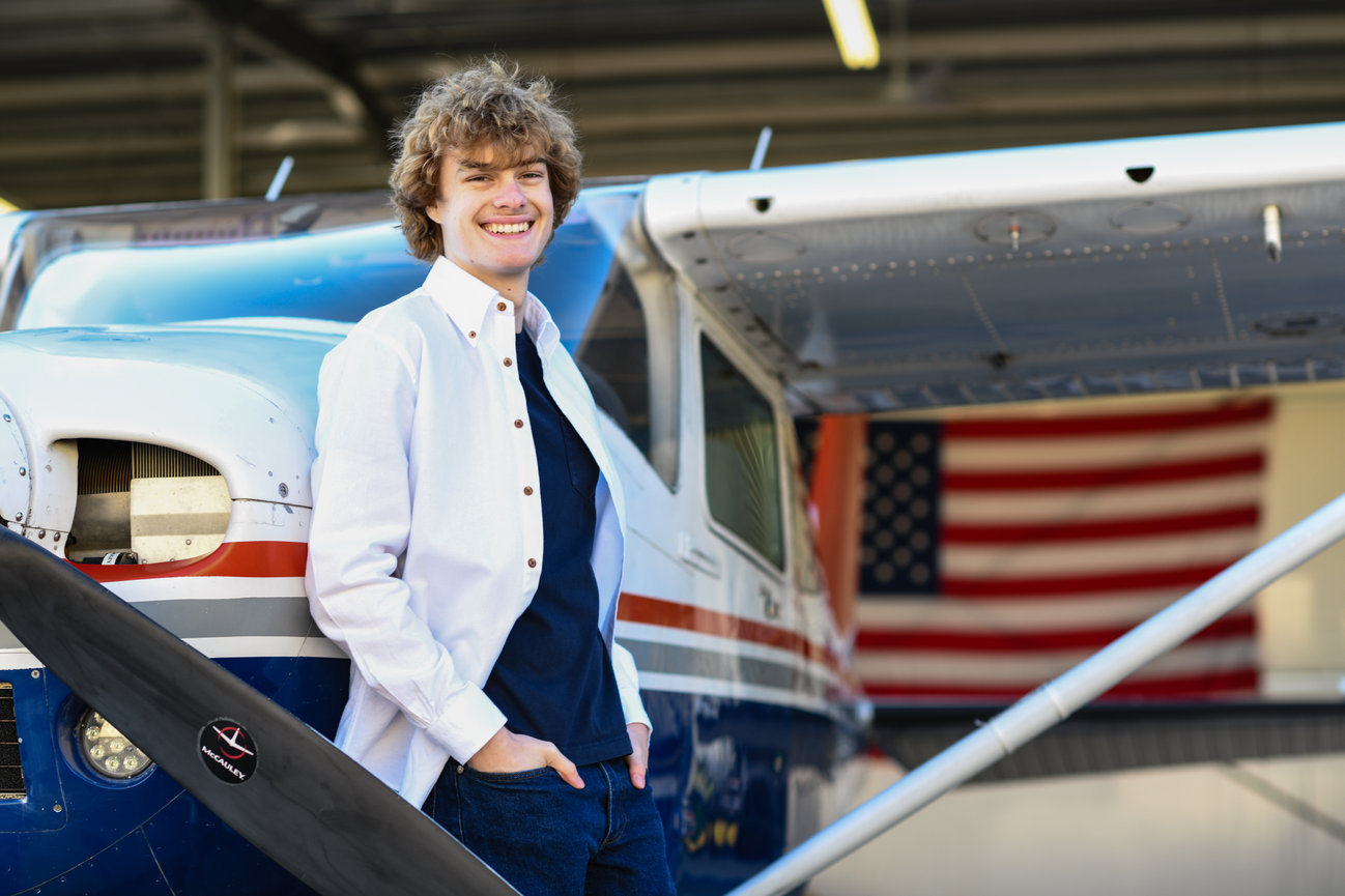Smiling young man in front of a vintage airplane, with an American flag in the background.