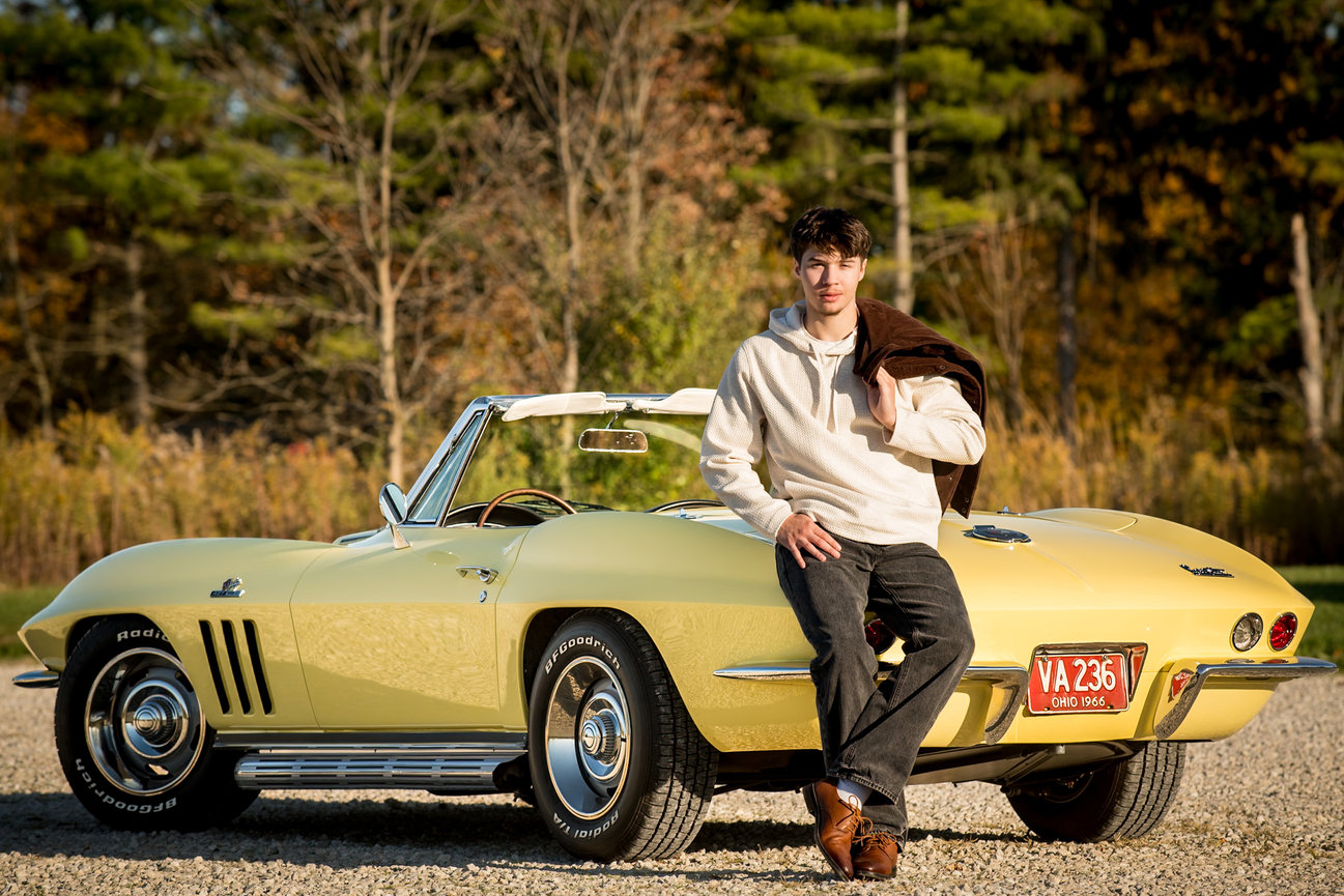 Teenage boy leaning against a classic yellow corvette in a fall colored wooded area. Photographed in Dublin Ohio for Senior Pictures. low convertible in a scenic outdoor setting.