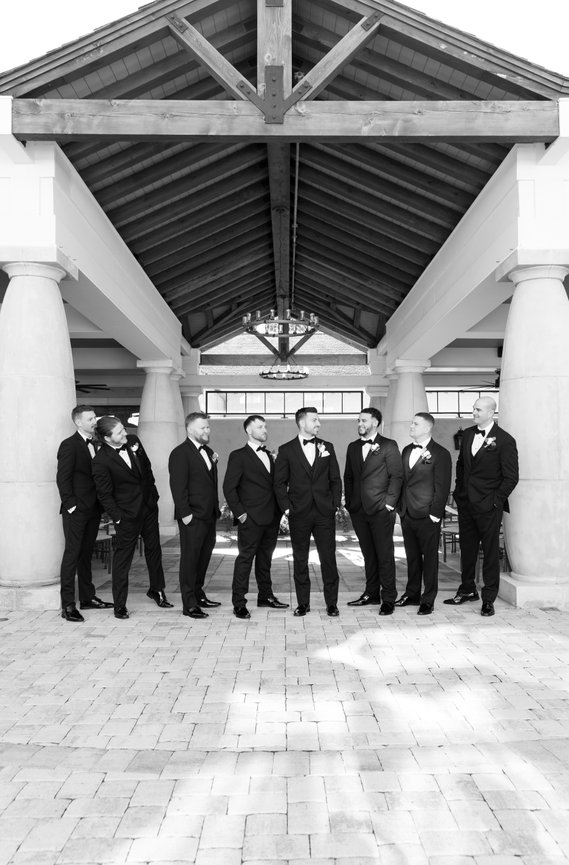 Groom and groomsmen in tuxedos stand in a line under a pavilion, smiling and talking.