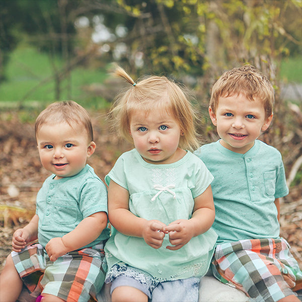 Three young children sit outdoors in matching teal shirts. The scene includes greenery in the background.