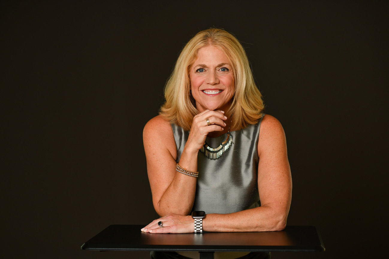 Blonde woman smiling, sitting at a table, wearing a gray sleeveless top and necklace against a dark background.