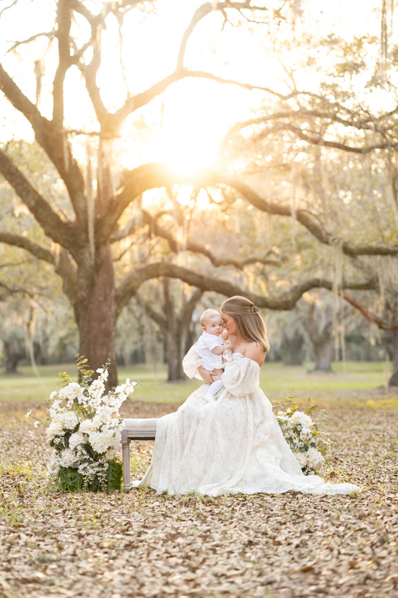 Mother in a white dress holding a baby outdoors at sunset with floral arrangements