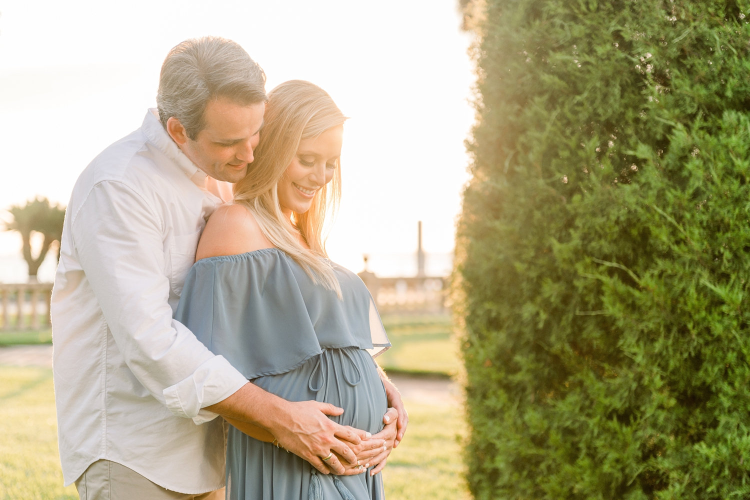 father and mother to be smiling and holding her pregnant belly, manicured tree in the right half of the photo