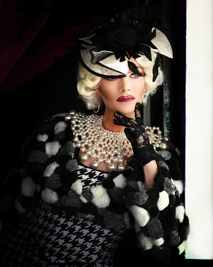 A glamorous drag performer poses by a window, wearing a stylish hat and pearl accessories.
