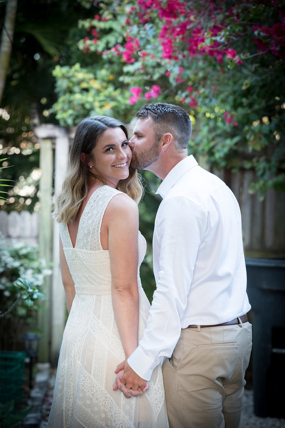 Couple in a garden setting, smiling and holding hands, with one partner kissing the other’s cheek.