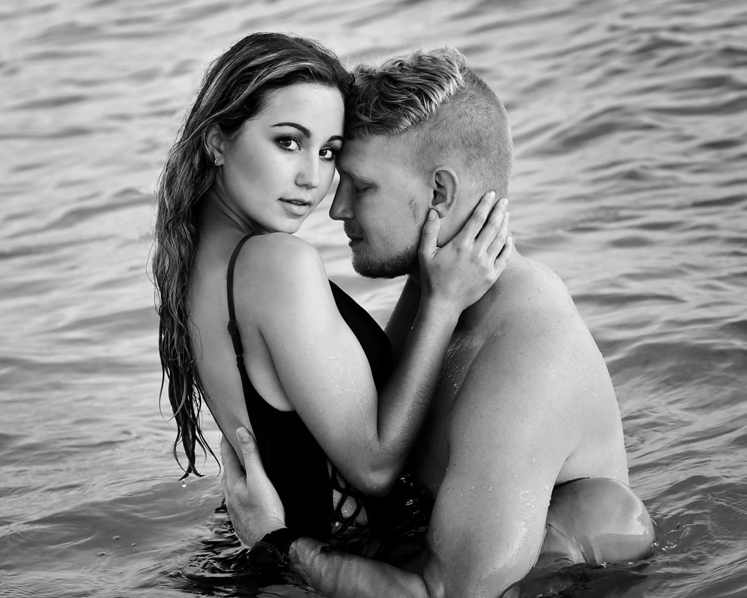 A couple embraces in a serene, black-and-white beach setting, with water gently surrounding them.