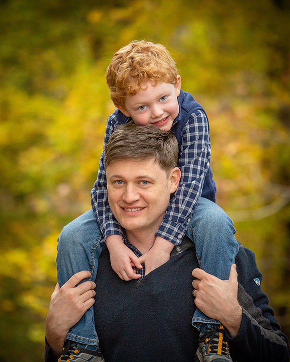 Man carrying smiling child on shoulders, surrounded by a blurred autumn forest background.