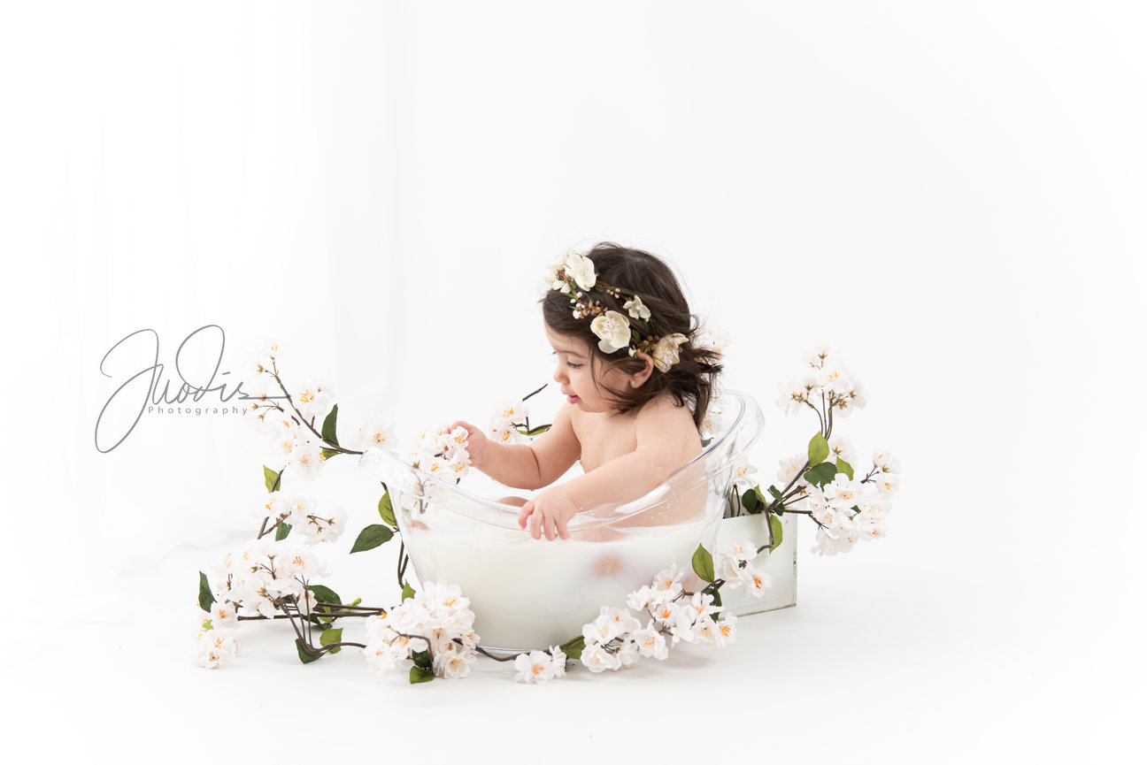 A young child sits in a clear tub filled with milk, surrounded by white flowers, against a light background.