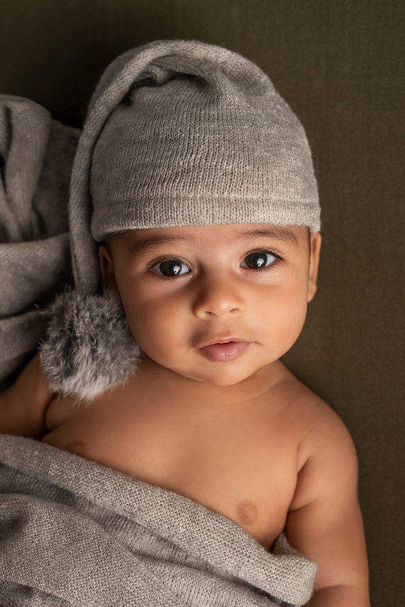 A baby with big eyes lies on a textured, dark green surface, wearing a soft, gray knitted hat with a pom-pom. Partially wrapped in a cozy matching gray blanket, this serene baby photos capture the little one gazing upward with a calm expression.